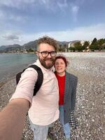 couple takes a cheerful selfie on a pebbled beach, with vast mountains and a serene sea in the background. their relaxed expressions and casual attire complement the picturesque coastal scenery photo