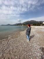 Millennial woman stands alone on a pebbled beach, gazing out at the tranquil sea under a partly cloudy sky. the serene setting is framed by distant mountains and a coastal cityscape photo
