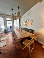 Cozy dining room with a long wooden table is elegantly set with wine glasses and natural decor. wicker chairs and pendant lights create a warm and inviting atmosphere, perfect for a gathering photo