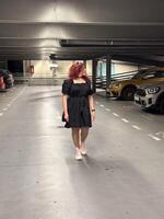 Millennial woman in a black dress walks through an empty parking garage, her pink sneakers adding a pop of color. Setting is urban and modern, with cars lined up neatly in the dimly lit space photo