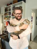 Warm moment between a millennial man and his cat, bond and affection shared in a cozy home environment. The background features a bookshelf at home. Pet and friendship concept photo