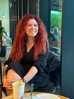 Millennial woman with red hair sits thoughtfully by a large window, gazing outside. Cozy cafe atmosphere, warm and serene mood. photo