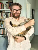 Warm moment between a millennial man and his cat, bond and affection shared in a cozy home environment. The background features a bookshelf at home. Pet and friendship concept photo