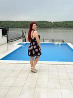 Woman with red hair in a black dress stands by a poolside, smiling and looking back at the camera. Happy vacation moment with a lush green river bank in background photo