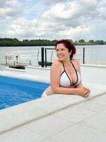Millennial woman with red hair relaxes in a pool on a calm day, looking thoughtfully into the distance. she rests her arms on the edge of the pool, enjoying the tranquil atmosphere and outdoor view. photo