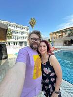 Smiling couple takes a cheerful selfie by a hotel pool on a sunny day, with mountains and clear skies in the background. Man and woman wearing casual outfits and joyful expressions photo