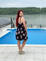 Woman with red hair in a black dress stands by a poolside, smiling and looking back at the camera. Happy vacation moment with a lush green river bank in background photo