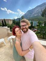 Millennial couple enjoys a vacation with a beautiful mountain backdrop while smiling and embracing on a sunny day taking selfie. Terrace photo