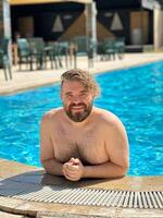 Millennial man enjoying a refreshing day by swimming pool, leaning on the pool's edge and beaming with a broad smile. Holidays and vacation concept photo
