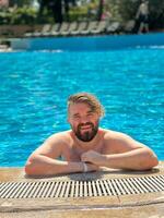 Man enjoying a refreshing day by swimming pool, leaning on the pool's edge and beaming with a broad smile. Blue water and relaxed setting create perfect atmosphere for summer fun. Holidays photo
