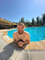 Millennial man enjoying a refreshing day by swimming pool, leaning on the pool's edge and beaming with a broad smile. Holidays and vacation concept photo