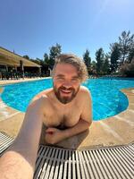 Millennial man taking selfie by swimming pool. Vibrant blue water and relaxed setting create a perfect atmosphere for summer fun. Holidays and vacation concept photo