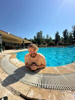 Millennial man enjoying a refreshing day by swimming pool, leaning on the pool's edge and beaming with a broad smile. Relaxed setting create a perfect atmosphere for summer fun. Holidays and vacation photo