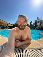 Millennial man taking selfie by swimming pool. Vibrant blue water and relaxed setting create a perfect atmosphere for summer fun. Holidays and vacation concept photo