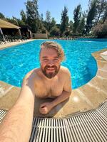 Millennial man taking selfie by swimming pool. Vibrant blue water and relaxed setting create a perfect atmosphere for summer fun. Holidays and vacation concept photo