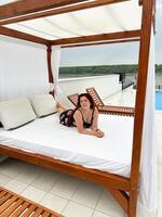 Millennial woman relaxing on a sunbed by a swimming pool, enjoying a serene view. Vacation tranquil setting beach bed and a scenic backdrop of lush greenery. photo