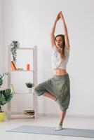 Woman practise yoga performs. Tree pose standing on mat inside of cozy room with plants and greenery. Healthy lifestyle concept. Copy space. photo