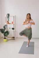 Woman practise yoga performs. Tree pose standing on mat inside of cozy room with plants and greenery. Healthy lifestyle concept. Copy space. photo