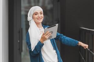 Smiling muslim girl using digital tablet, browsing Internet or social networks outdoor photo