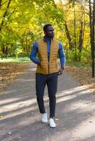 Cheerful african american young man in stylish clothes walks in autumn park on sunny warm autumn day. Concept of weekend and outdoor recreation photo