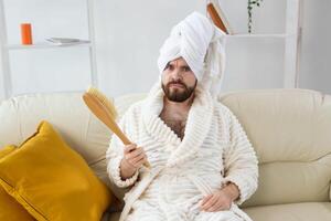 Portrait of cheerful guy wear turban towel holding in hands massage brush while sits on sofa. Male skin care and spa concept. photo