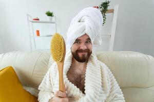 Portrait of funny guy wear turban towel holding in hands massage brush while sits on sofa. Male skin care and spa concept. photo