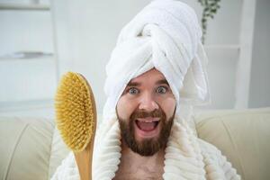 Portrait of funny guy wear turban towel holding in hands massage brush while sits on sofa. Male skin care and spa concept. photo