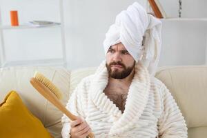 Portrait of cheerful guy wear turban towel holding in hands massage brush while sits on sofa. Male skin care and spa concept. photo