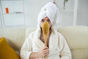 Portrait of funny guy wear turban towel holding in hands massage brush while sits on sofa. Male skin care and spa concept. photo