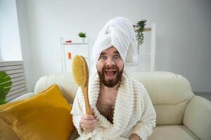 Portrait of funny guy wear turban towel holding in hands massage brush while sits on sofa. Male skin care and spa concept. photo