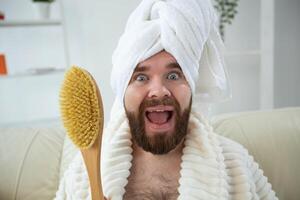 Portrait of funny guy wear turban towel holding in hands massage brush while sits on sofa. Male skin care and spa concept. photo
