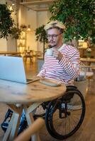 Focused guy freelancer with disabilities in wheelchair working online using laptop sits in cozy cafe photo