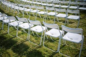 Folding chairs standing in row on green grass, casting shadow on ground. Outdoor party preparation photo