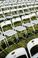 Folding chairs standing in row on green grass, casting shadow on ground. Outdoor party preparation photo