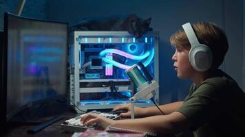 Young boy, wearing headphones and sitting in front of a microphone, enthusiastically and emotionally engages in computer shooting games at home in the evening. video