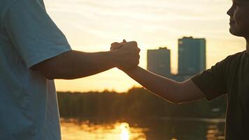 Firm handshake between the older and younger brother, with the backdrop of the setting sun illuminating the lake in the city park. video