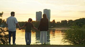 Mother, father, and son are standing against the backdrop of a setting sun in a city park, in front of a beautiful lake. They are holding hands and communicating with each other. video