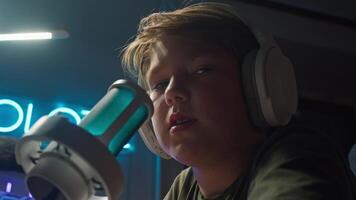 A teenage boy with red hair records his podcast using neon lighting, a professional microphone, and wireless headphones. Close up video