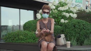 Ederly woman in medical mask sits on bench in city and disinfects her dog dachshund hands and paws with antiseptic. Senior female masked with pet use anitizer in park during quarantine covid 19. video