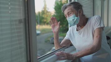 Portrait of masked senior woman in late 90s waving from home window. Elderly woman in protective mask looks out the window and waves her hand while isolating at home and conducting social distancing. video