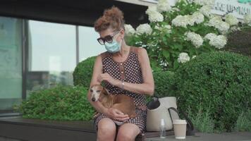 Senior woman in protective mask resting on bench in city with small dog Dachshund breed. Elderly female and pet on bench during quarantine coronavirus. Masked mature owner with dog sitting in park. video
