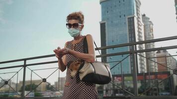 Elderly dog owner in medical mask uses an alcohol antiseptic while walking in the city. Covid time, new rules and restrictions. Mature female in mask disinfecting hands with pet in carrier outside. video