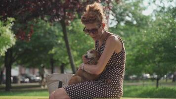 A mature woman sits on a bench in a city park with a dachshund dog holding it in her arms and hugging. Happy elderly woman sitting with dog on bench in summer park. Rest with a pet. video