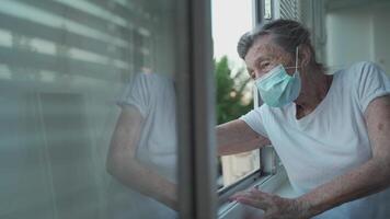 Portrait of masked senior woman in late 90s waving from home window. Elderly woman in protective mask looks out the window and waves her hand while isolating at home and conducting social distancing. video