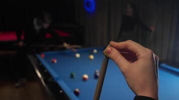 Woman's hand rubs the tip of a billiard cue with a special slip-preventing chalk. In the background, her opponent is playing in the American pool video
