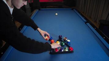 Young man puts multicolored billiard balls on blue American pool table in a triangle for placing balls in initial position of a pyramid shape video