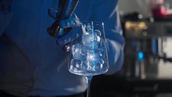 A barman in a nightclub serves a red martini, using a jigger, into a glass filled with ice that sparkles in the neon light. Close up video