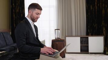 Young businessman working from hotel room on business trip. A man sitting on bed and using laptop video