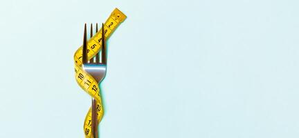 Close up of fork wrapped with tape measure on blue background. Top view of obesity concept photo
