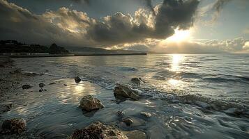 A dramatic sunset over a calm sea with large, dark clouds, the sun shining through and reflecting off the water. There are a few rocks in the foreground and a small wave is breaking on the beach. photo
